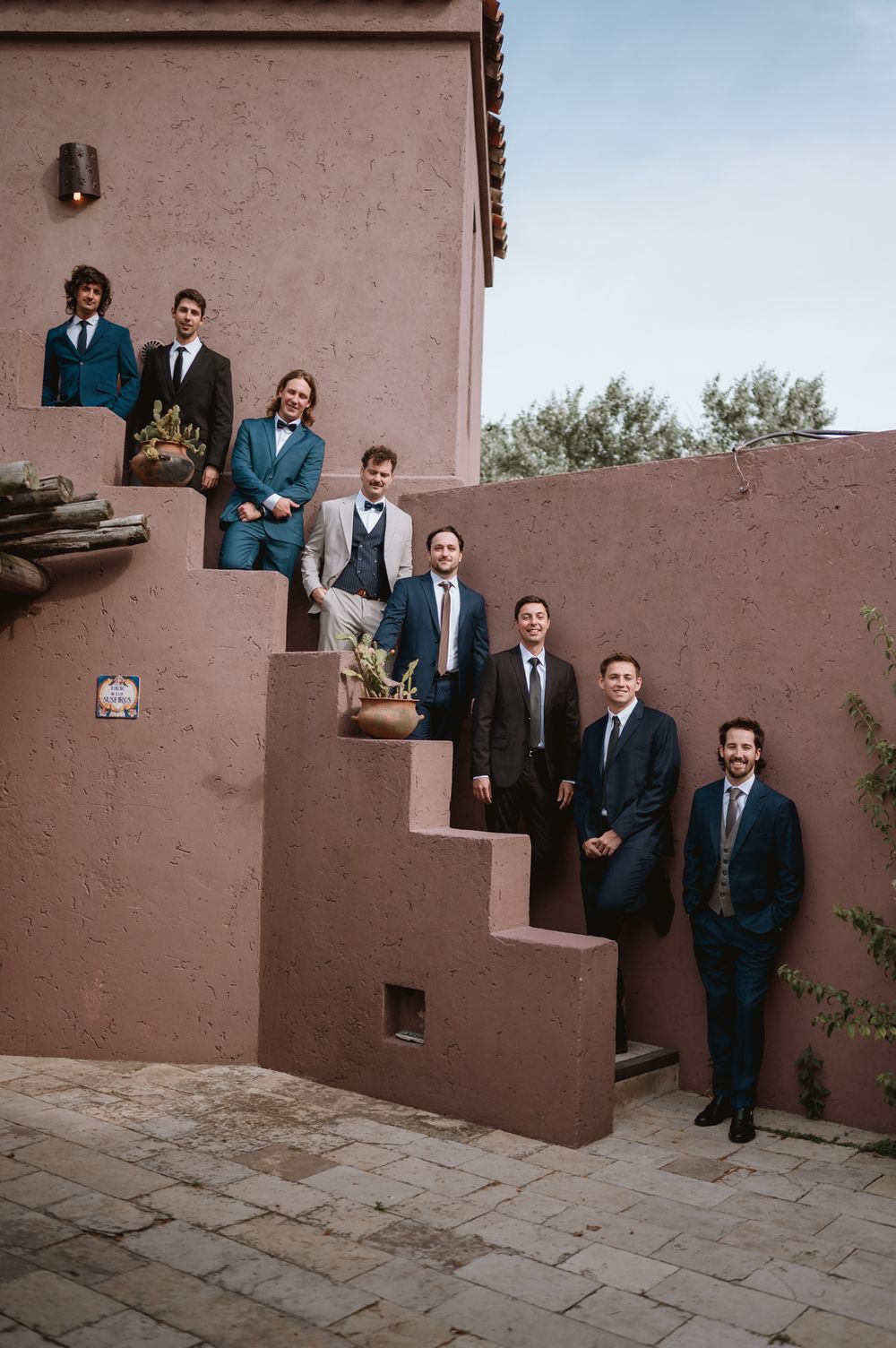 Groom and groomsmen posing on outdoor stairs of a pink stucco wedding venue.