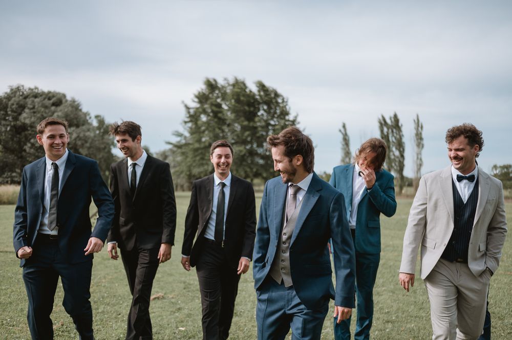 Groom and groomsmen laughing and walking together across a green field in suits.