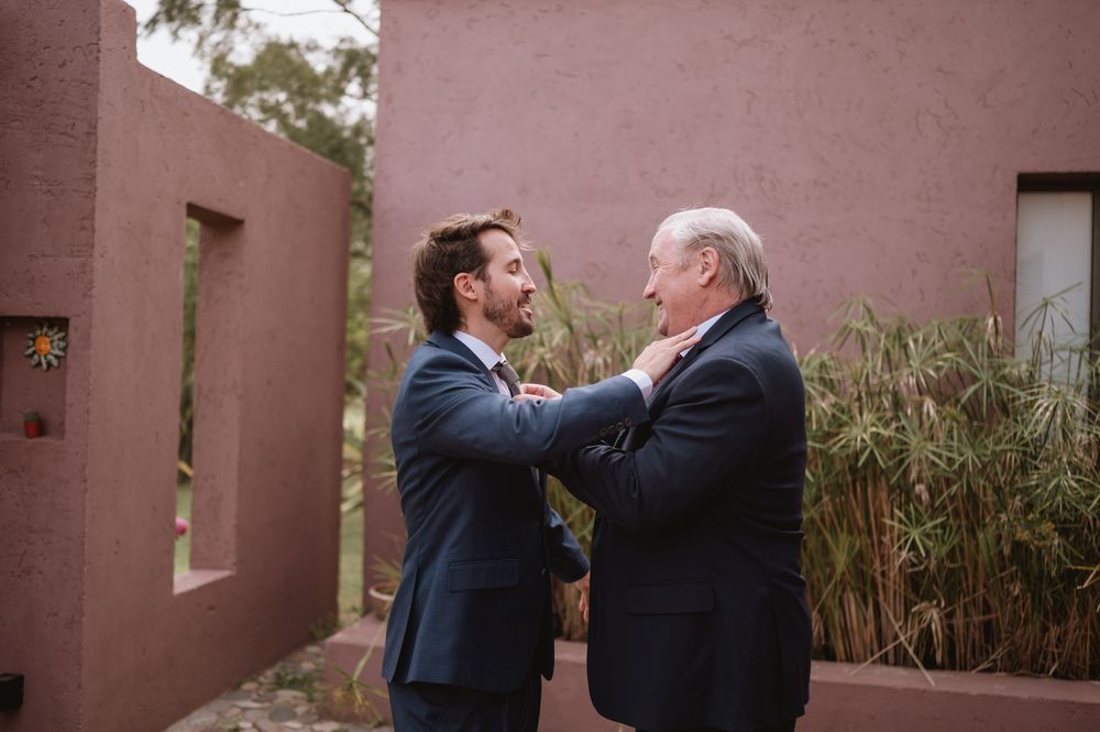 Groom adjusting older mans tie outdoors near pink stucco wall at wedding.