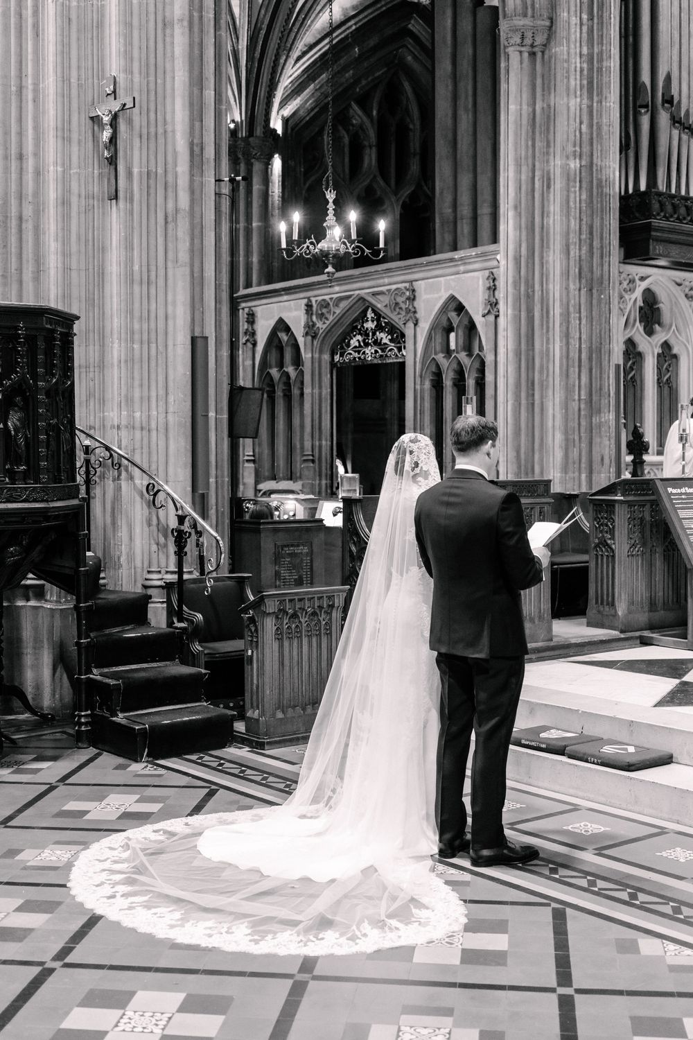 Bride and groom standing at cathedral altar, bride wearing long lace veil, black and white photo.