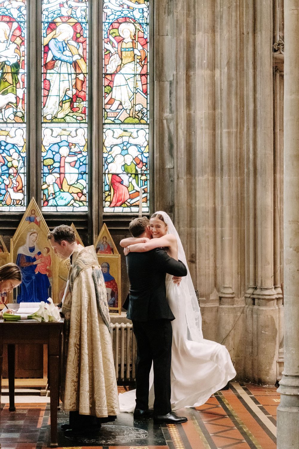 Bride and groom embrace before stained glass window as priest witnesses church wedding ceremony.