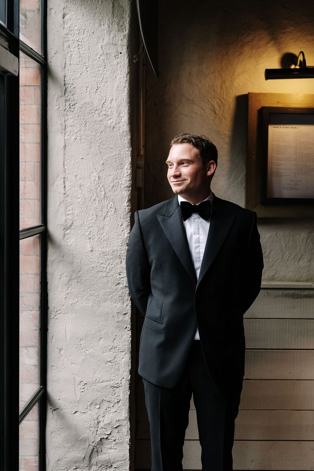 Groom in black tuxedo with bow tie standing by window in elegant venue, smiling softly. Pasture, St Mary Redcliffe Church, Bristol Wedding