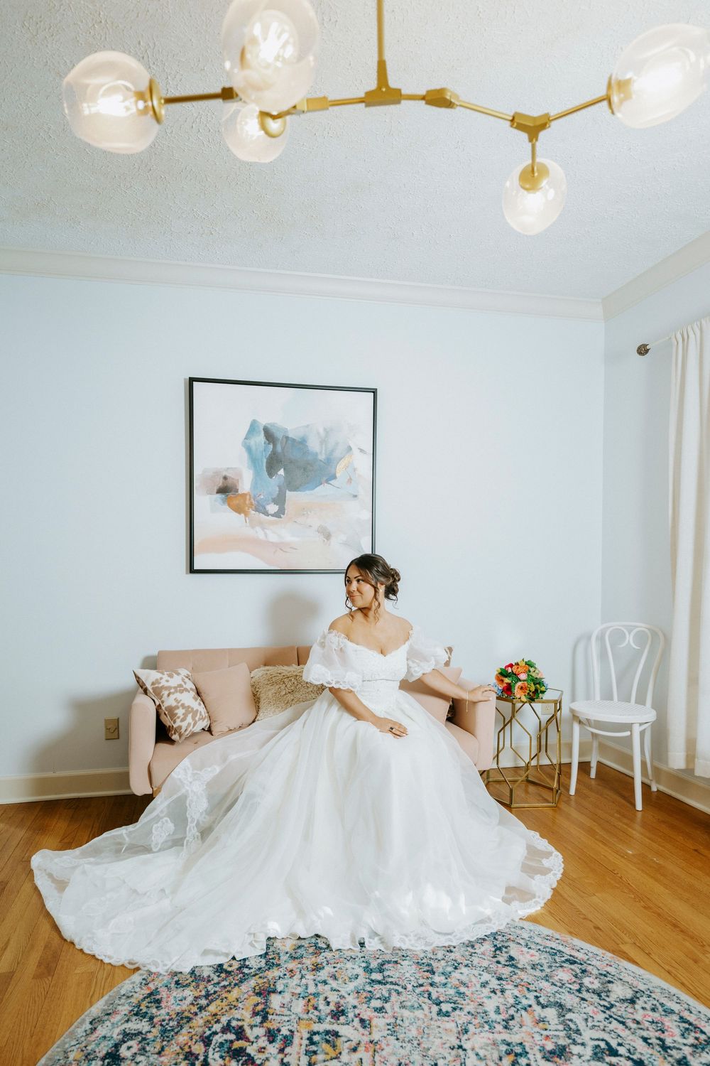 Bride in a white off-shoulder ball gown sitting in a bright room with floral rug and modern gold chandelier.