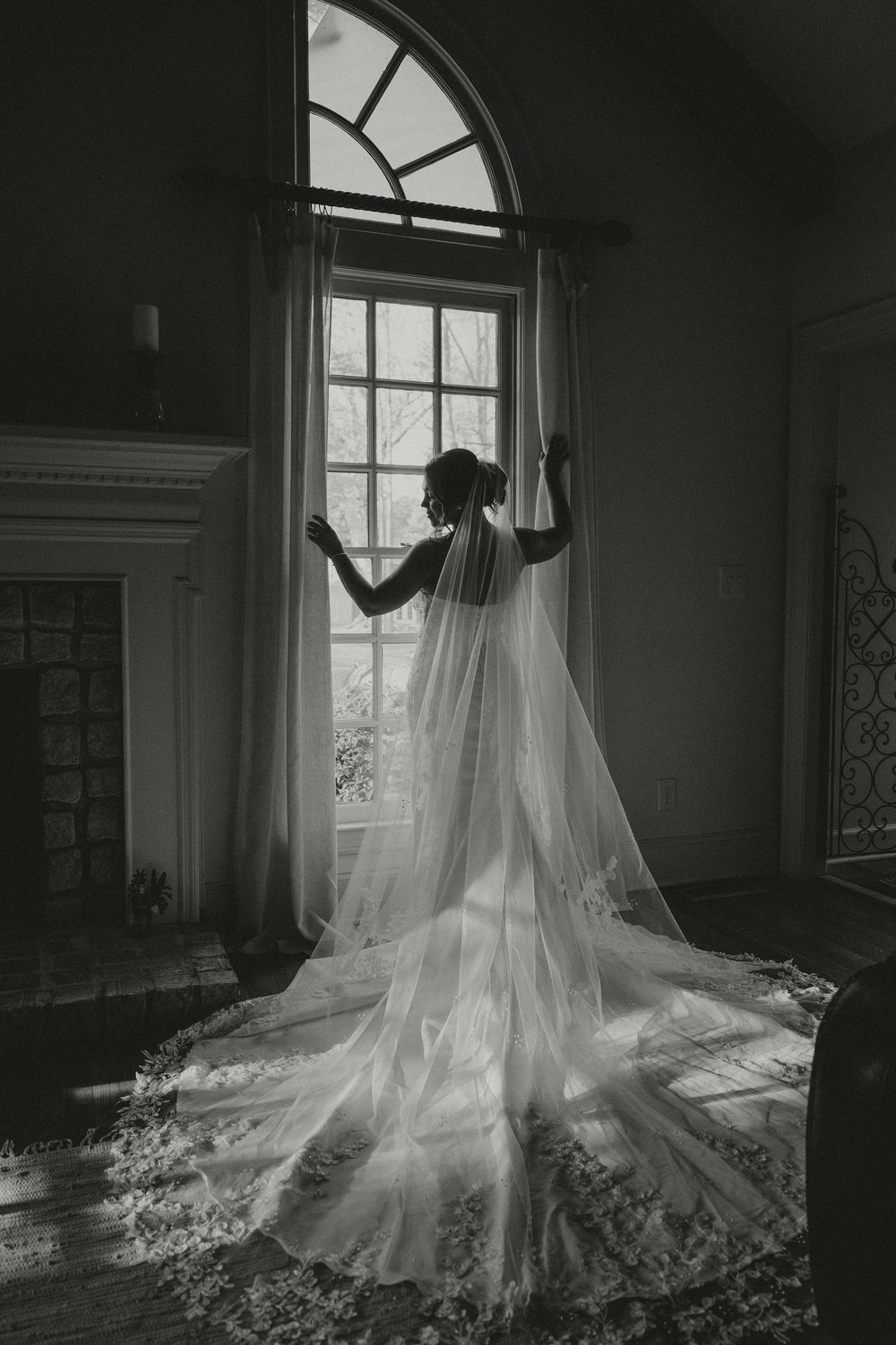 Black and white photo of a bride in a flowing veil standing before an arched window in an elegant room.