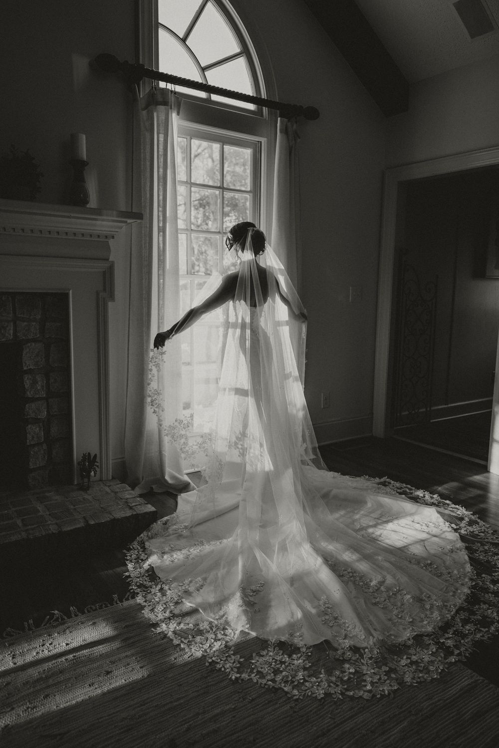 Black and white photo of a bride in a flowing gown and lace veil standing by a sunlit window in an elegant room.