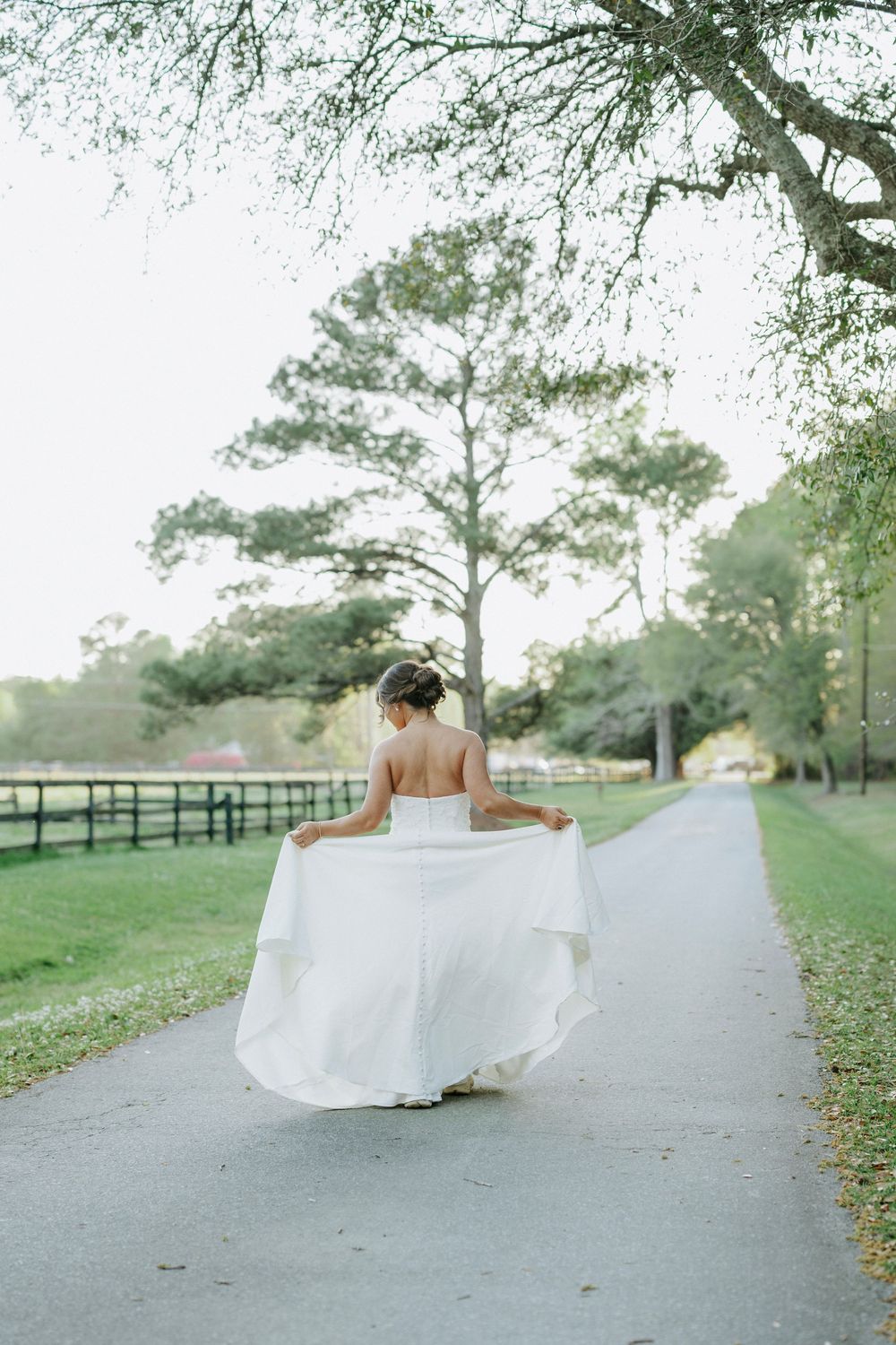 Bride in white gown walks down tree-lined path at outdoor wedding venue, holding her flowing dress train.