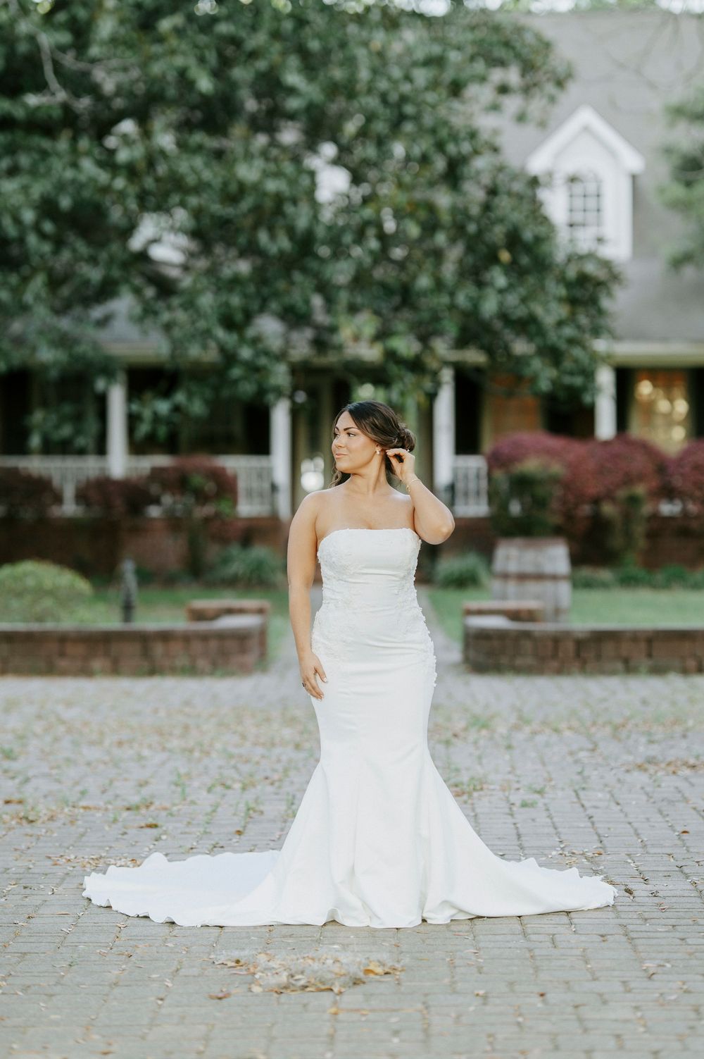 Bride in strapless fitted white wedding gown with train posing on brick courtyard before elegant estate.