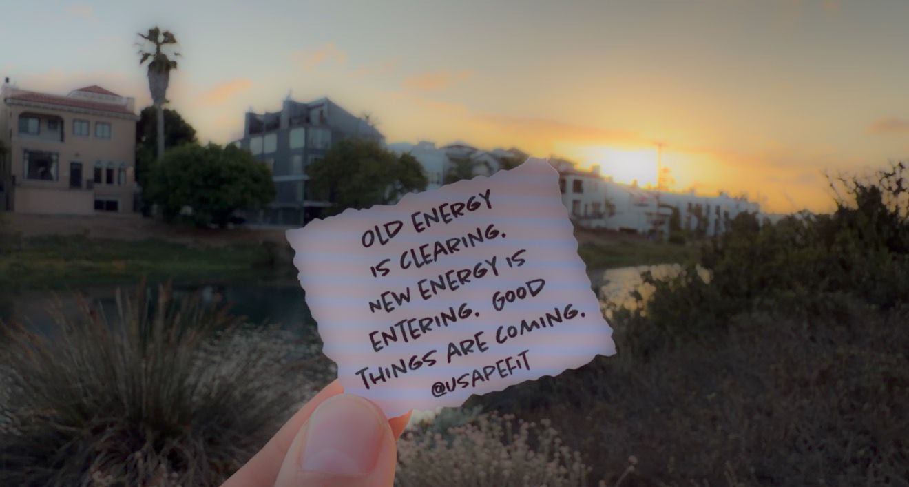 Hand holding a motivational note reading old energy is clearing new energy is entering good things are coming at sunset.