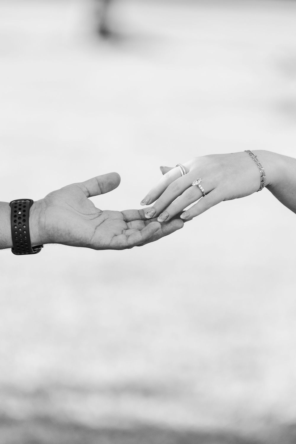 Black and white photo of two hands reaching toward each other, one wearing a ring and bracelet, the other a leather bracelet.