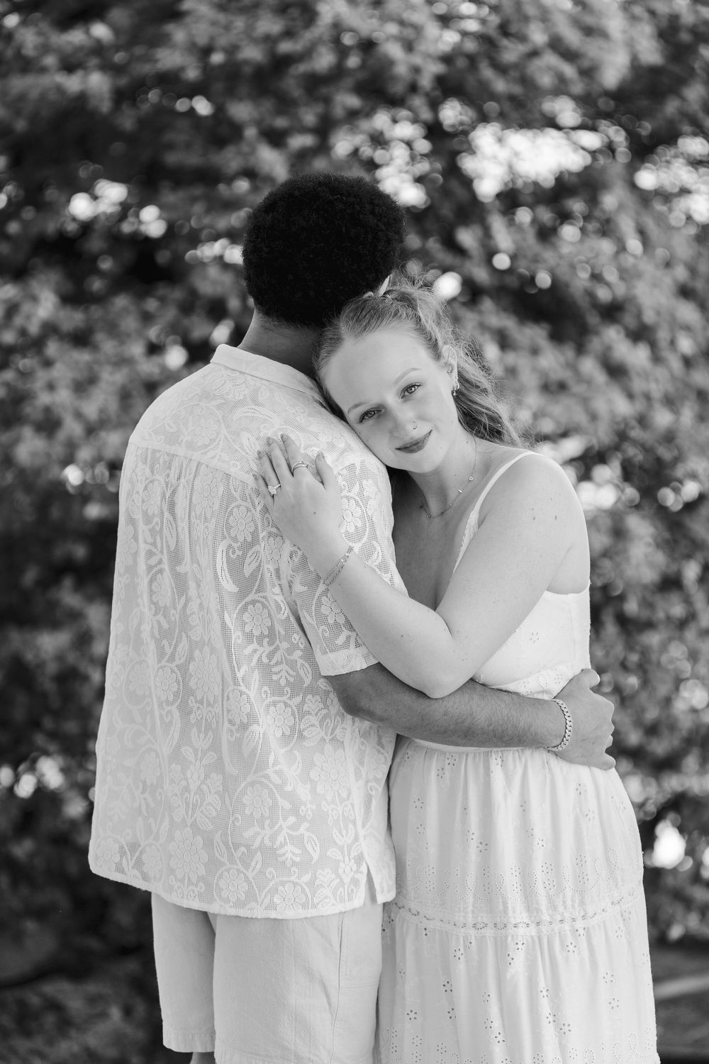 Black and white photo of an interracial couple embracing outdoors, woman in white dress resting head on mans shoulder.