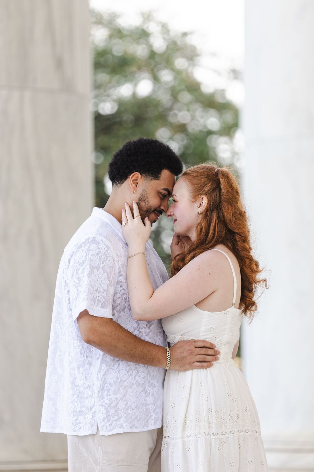 A romantic interracial couple in white outfits share a tender moment, touching foreheads between marble columns.