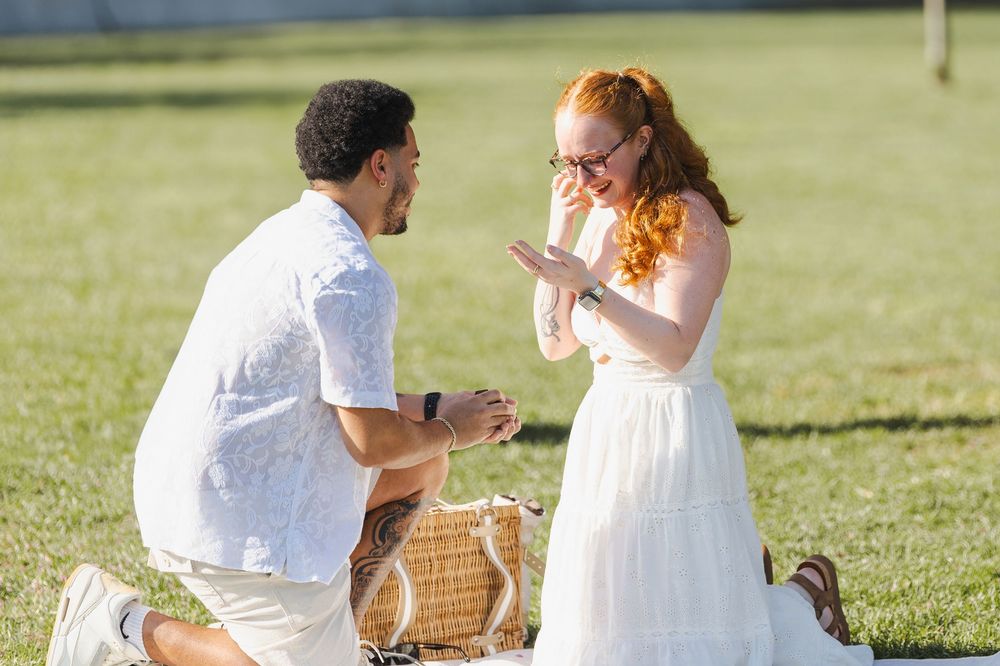Man proposing to a surprised red-haired woman on a picnic blanket in a sunny green park.