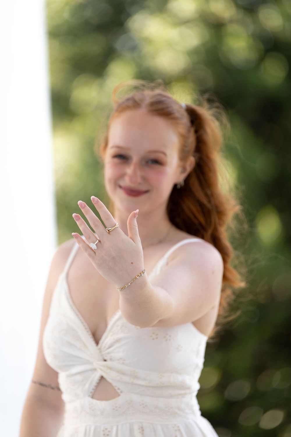 Young woman with red hair in white dress showing hand with rings and bracelet jewelry outdoors.