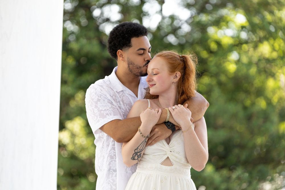 Romantic couple embracing outdoors, man kissing woman's temple near white column with lush green trees background.