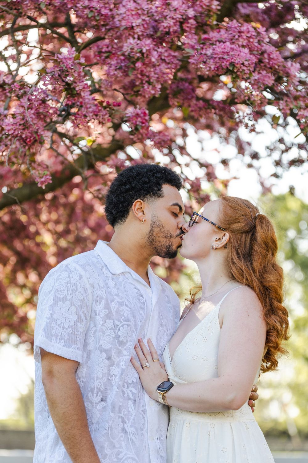Couple sharing a romantic kiss under a blooming pink cherry blossom tree in spring.