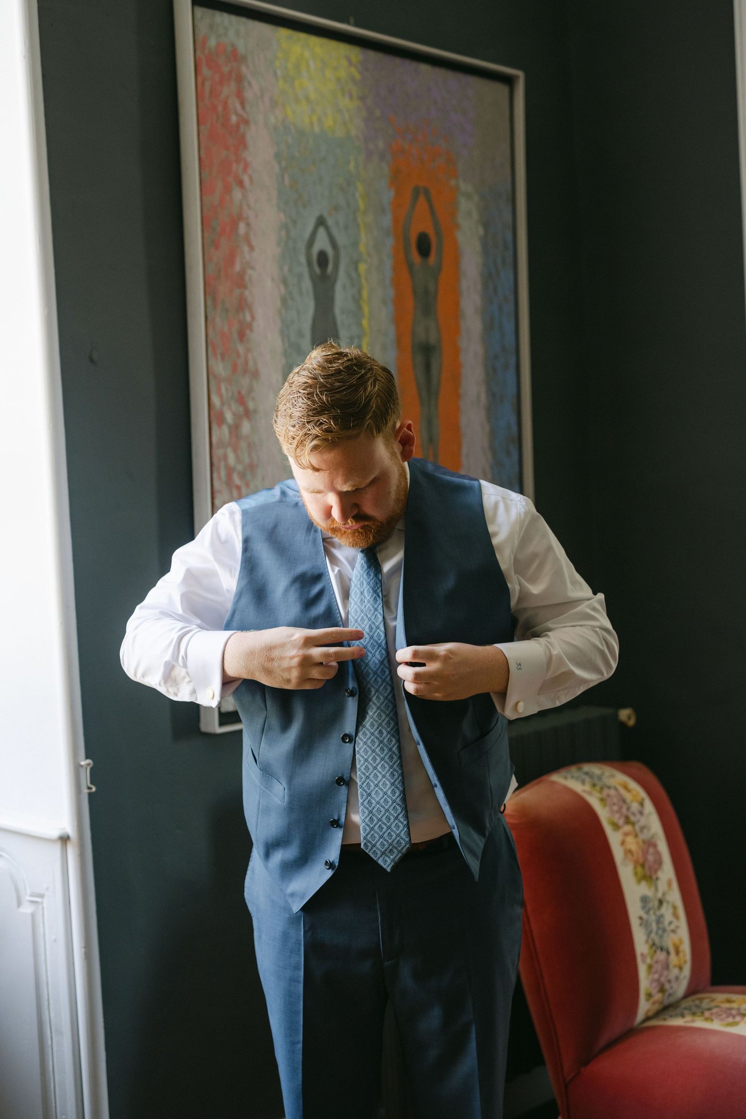 Groom in a blue three-piece suit stands confidently in a doorway of an elegant room with dark teal walls.