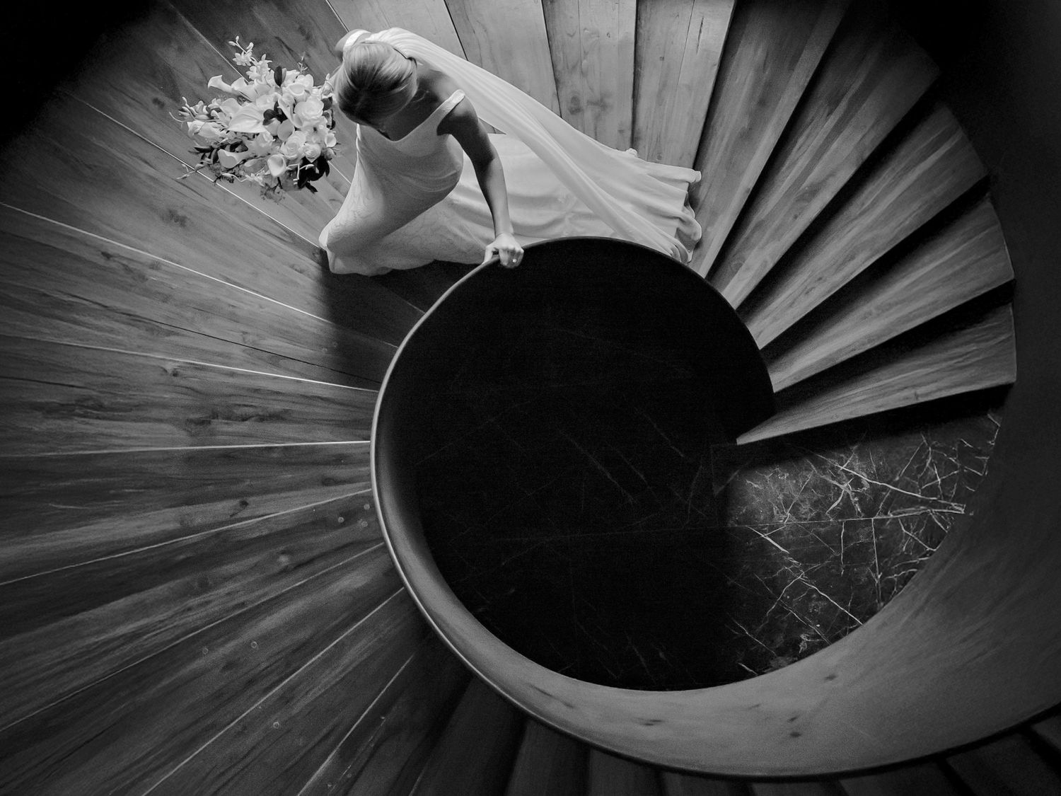 Black and white aerial view of a bride in a white gown descending an elegant spiral staircase holding a bouquet.