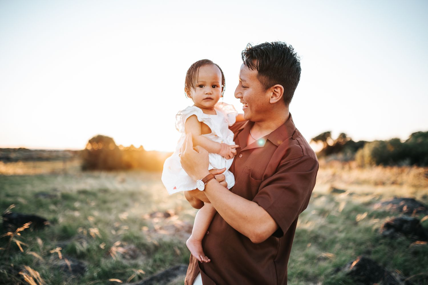 A father holds his baby girl in a sunlit field at golden hour, sharing a tender moment outdoors taken by Melbourne family photographer.
