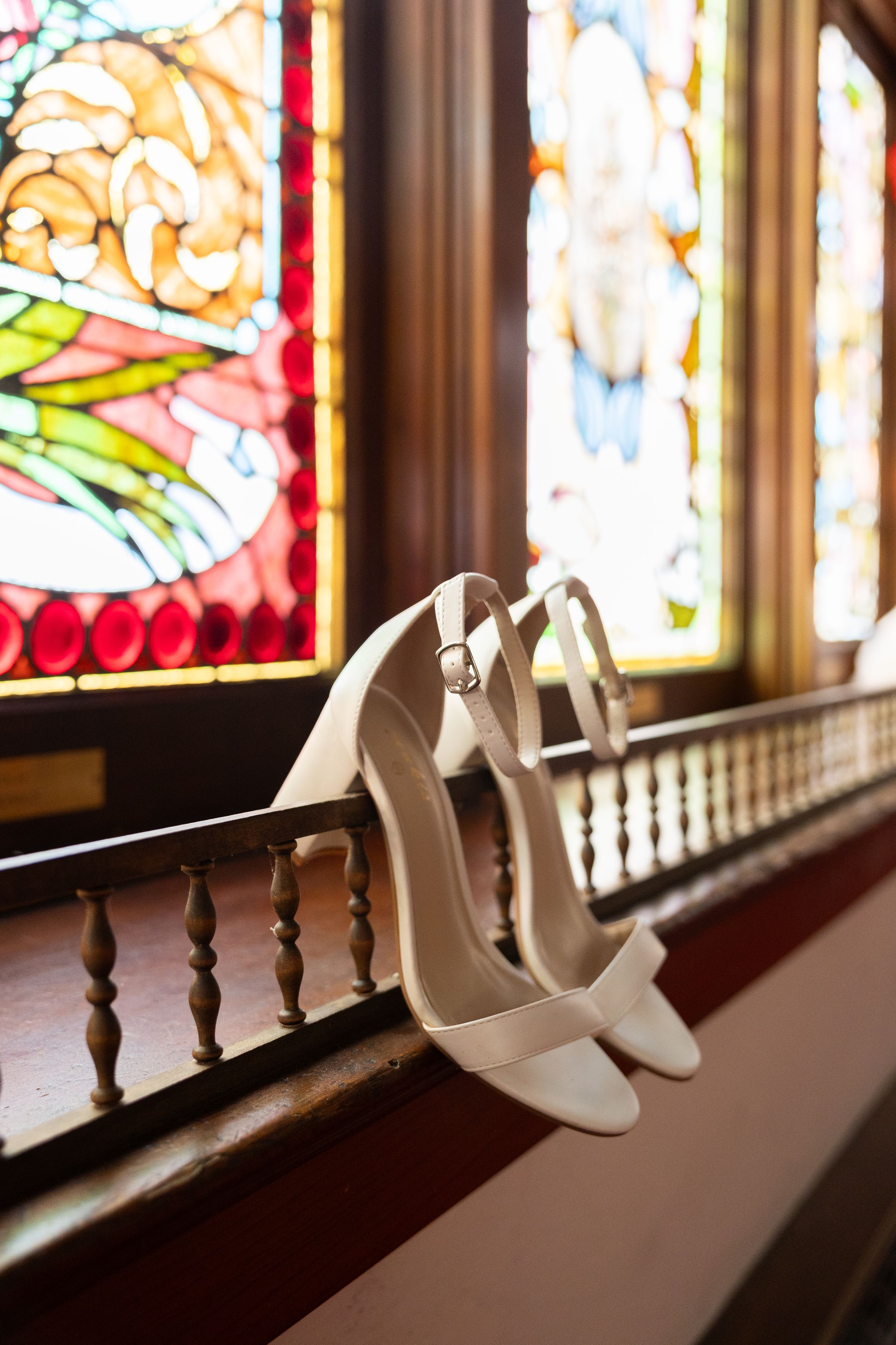 White bridal heels resting on a wooden railing in front of colorful stained glass windows in a church.