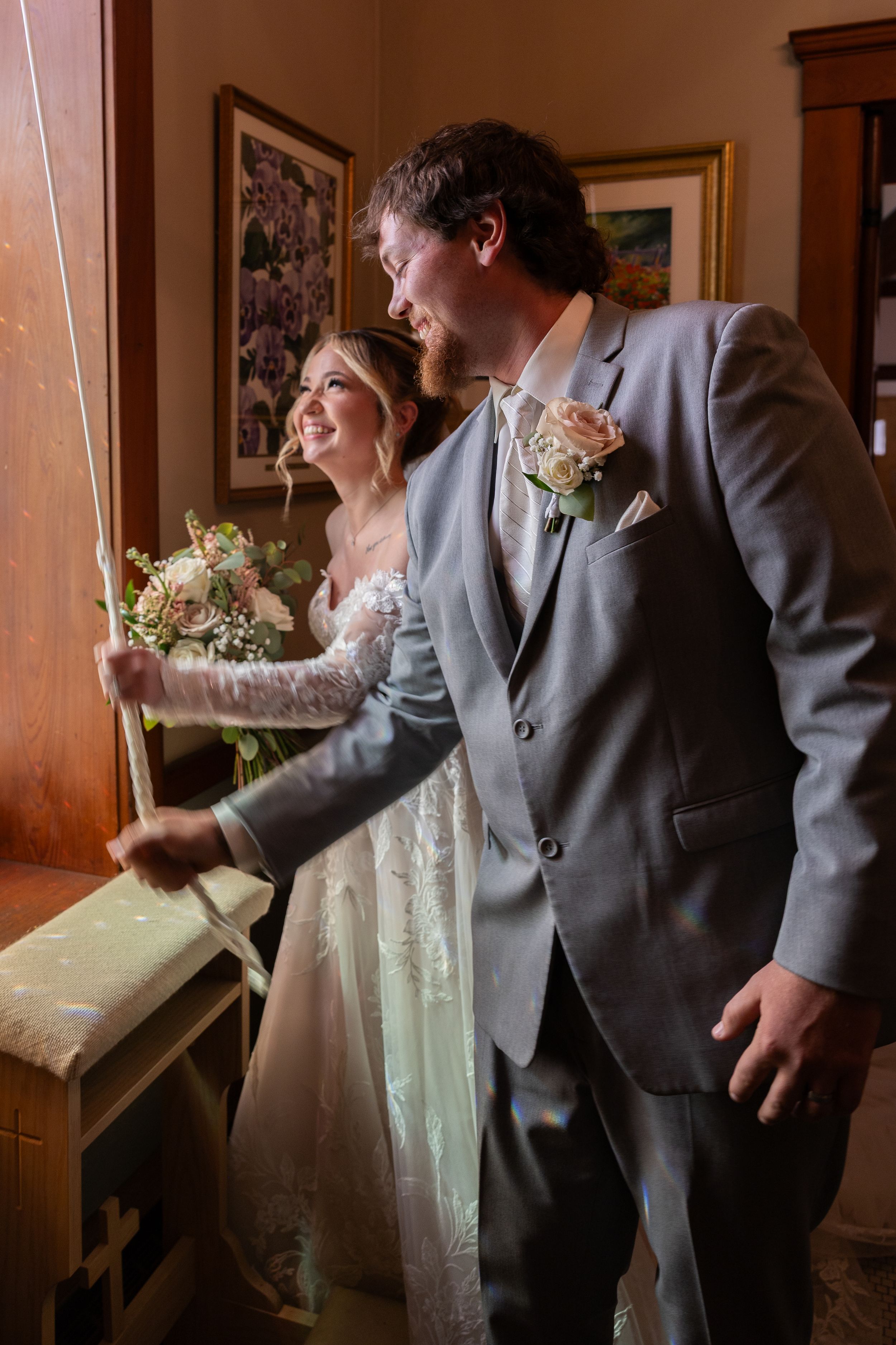 Bride and groom laughing together indoors, bride holding a bouquet, groom in gray suit with boutonniere.