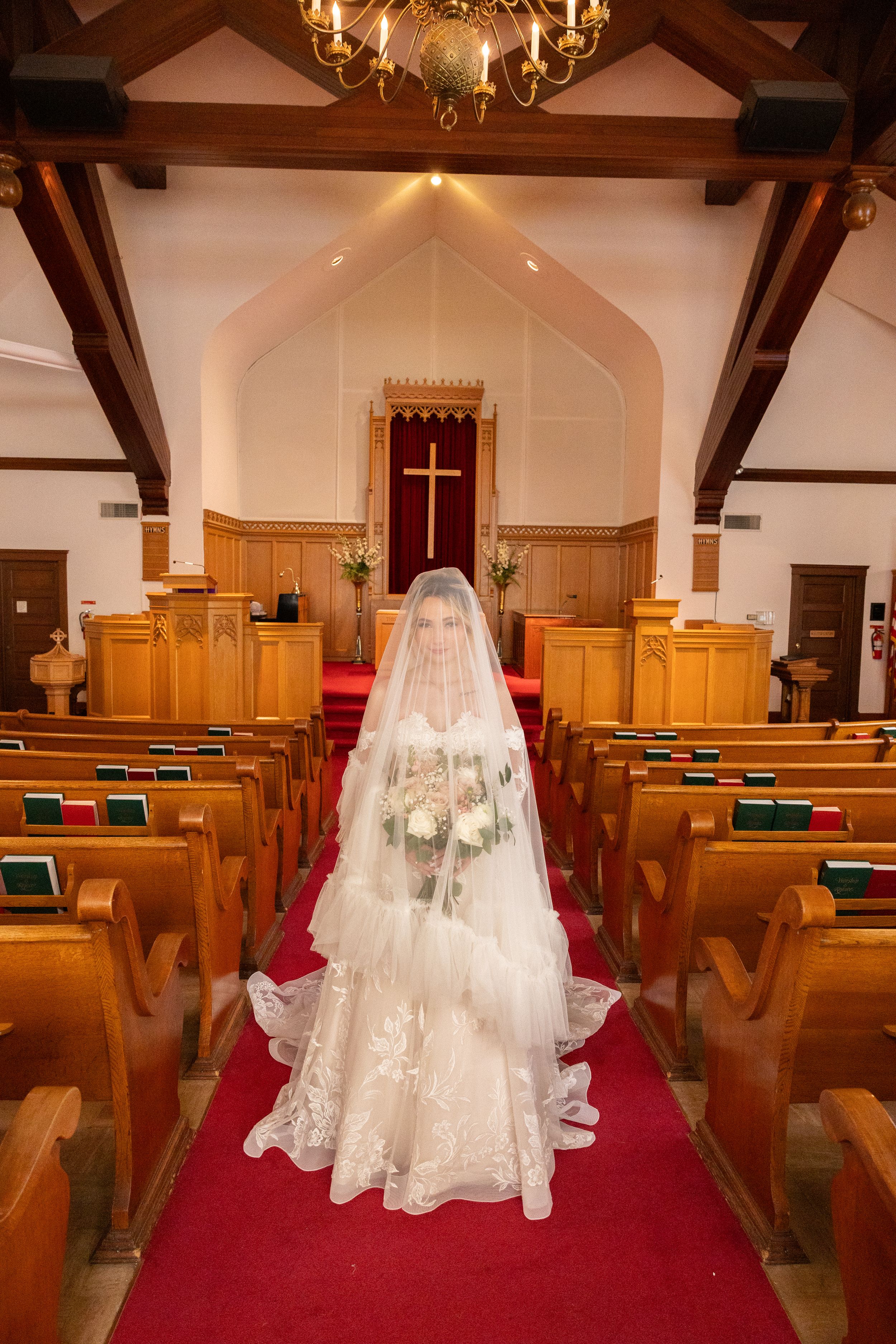 Bride in lace gown and veil stands on red carpet aisle of traditional church holding white bouquet.