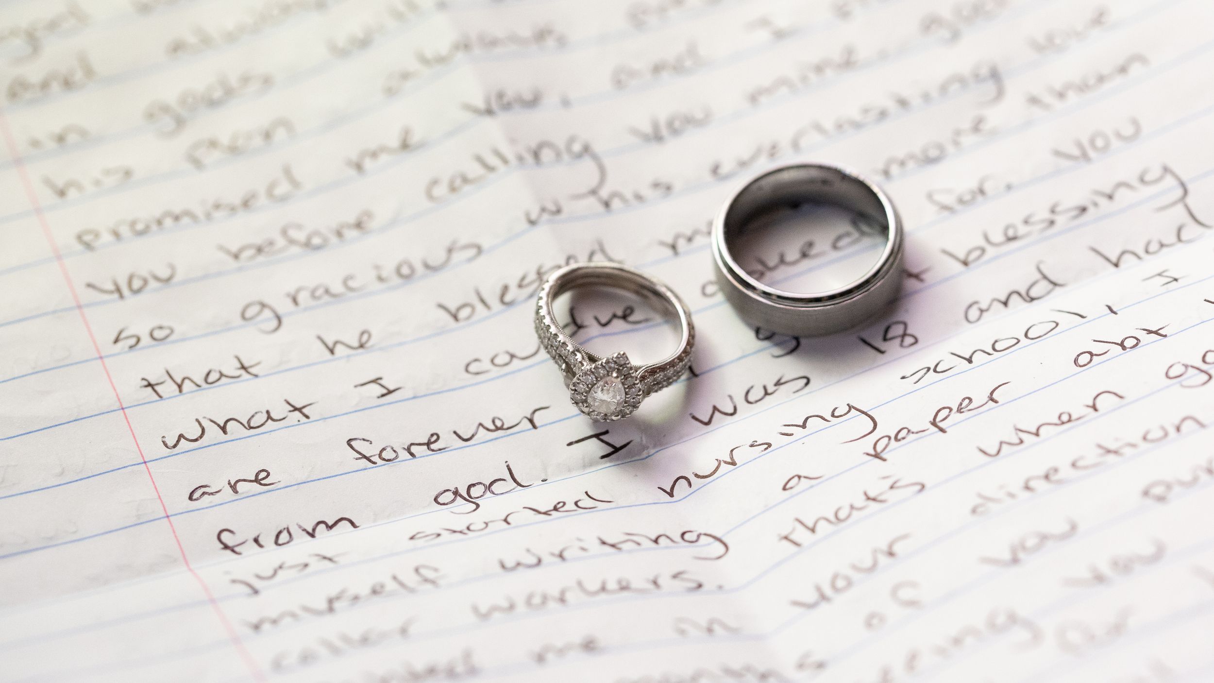 Wedding rings resting on handwritten vows letter, diamond engagement ring and silver band on paper.