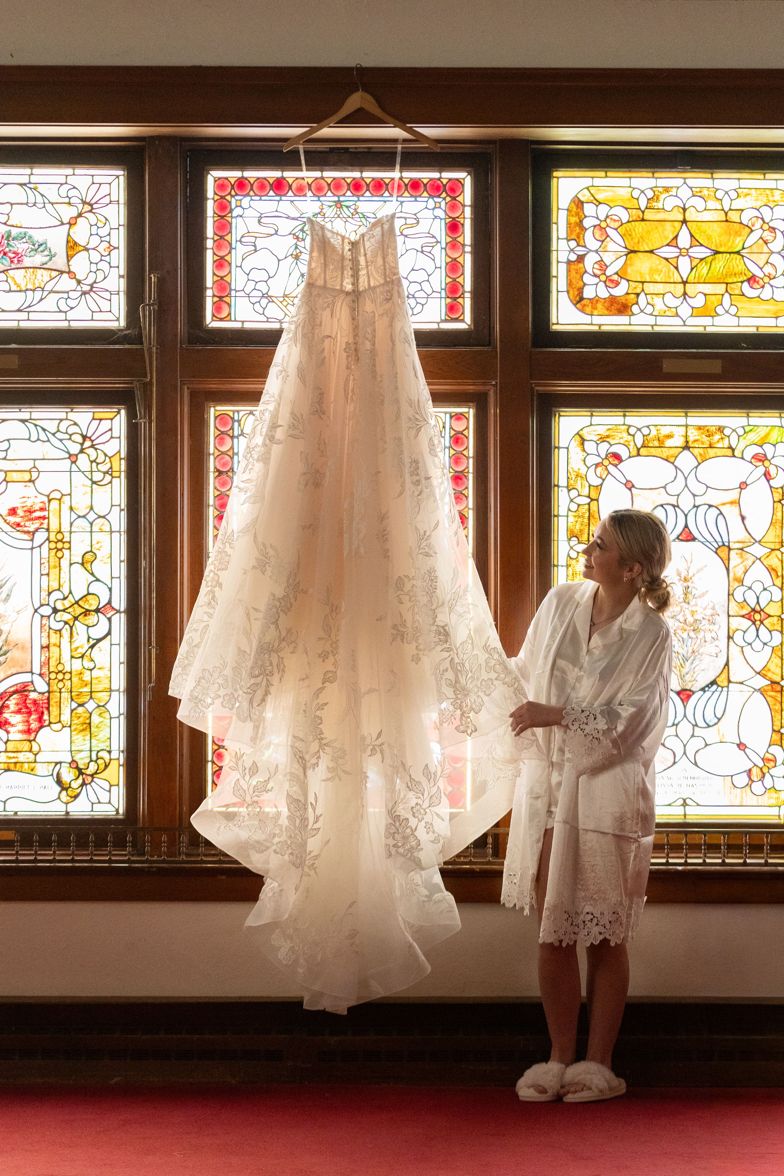 Bride in white robe admires lace wedding dress hanging before colorful stained glass church windows.