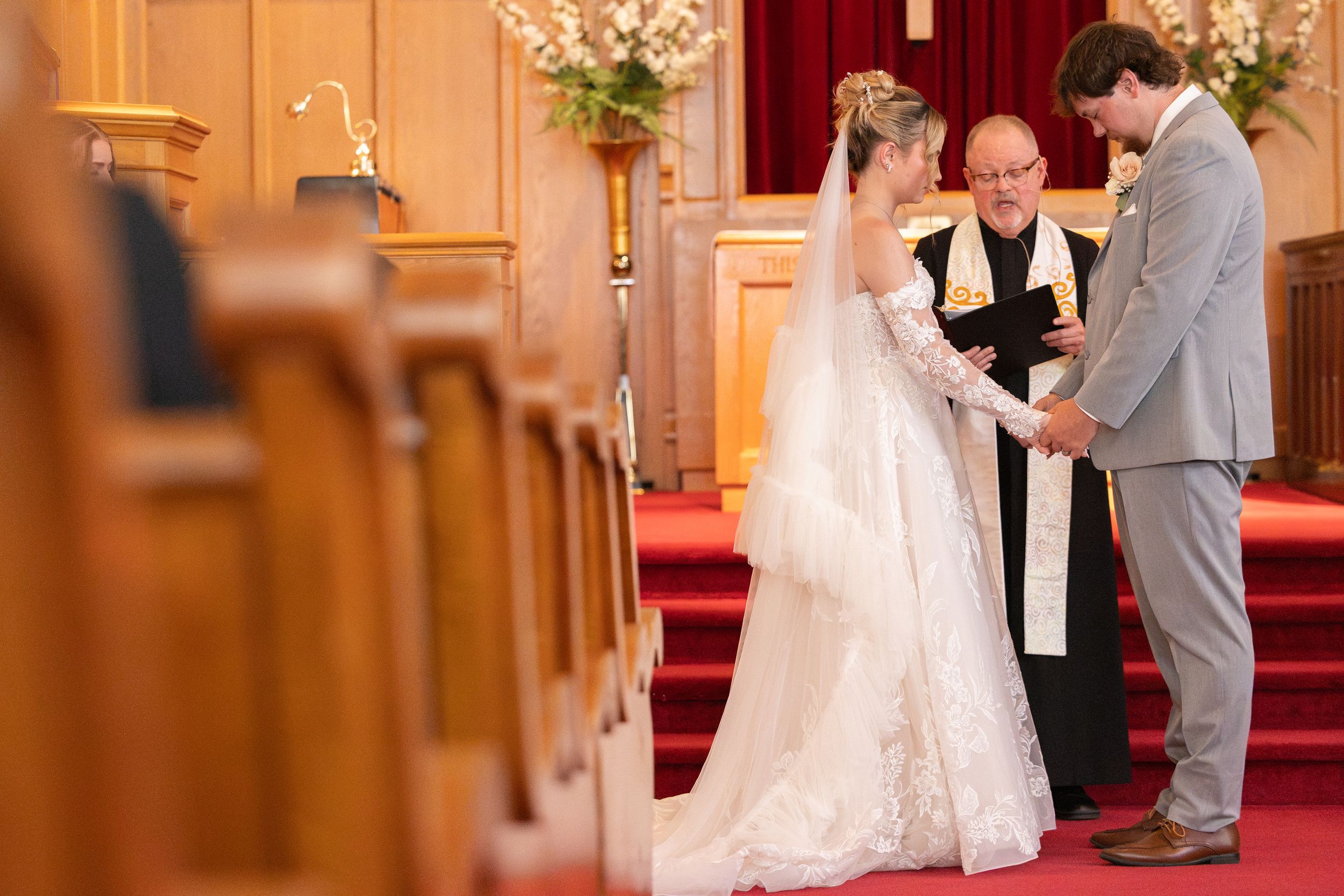 Bride and groom exchanging vows with officiant at church altar, bride in lace gown, red carpet aisle, wooden pews.