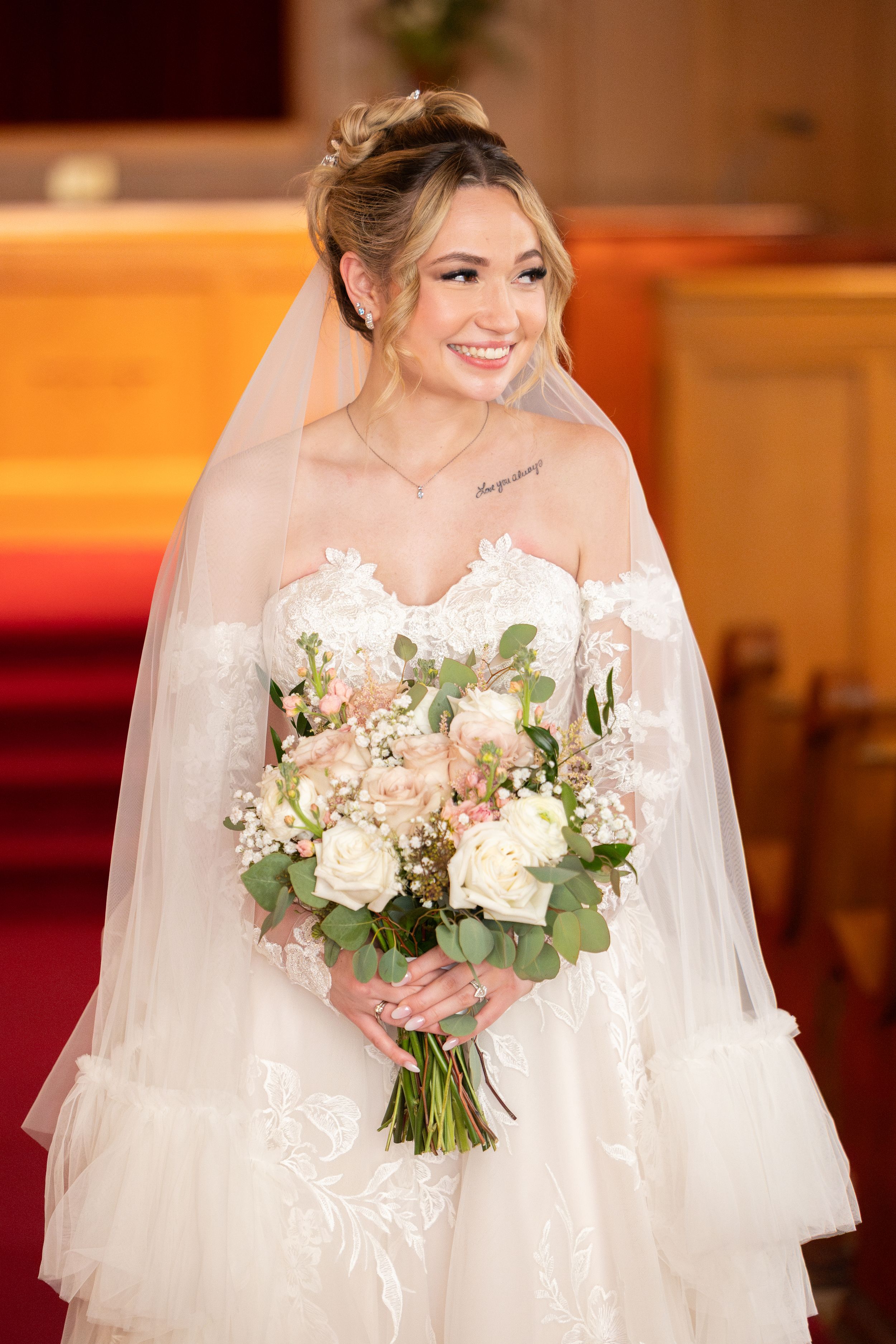 Smiling bride in strapless lace gown and veil holds white and blush bouquet inside a church.