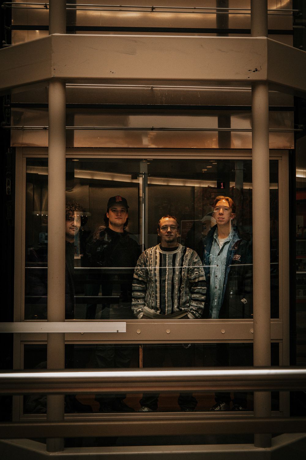 Three people standing inside a glass elevator at night, lit dramatically from below, wearing casual streetwear.