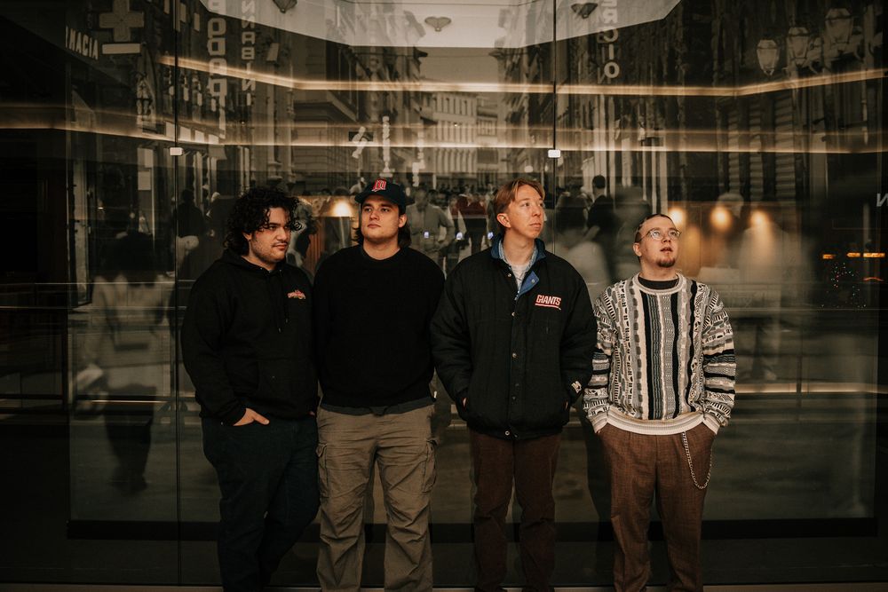Four young men standing together in front of a reflective glass building facade in an urban setting.
