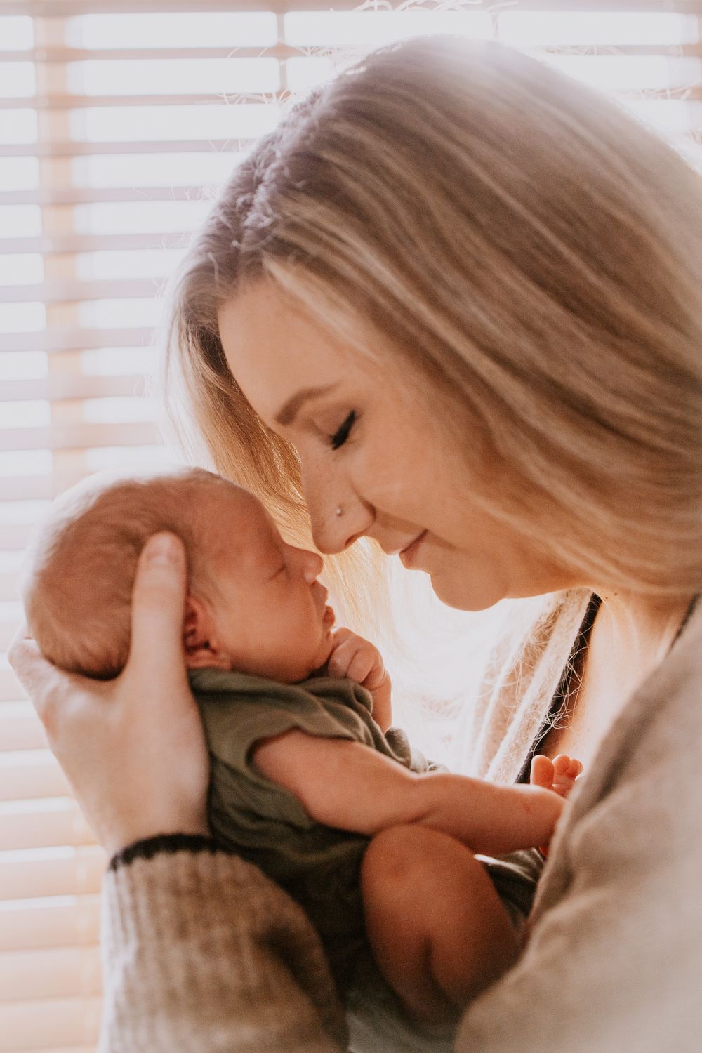 A tender moment as a young blonde mother gazes lovingly at her newborn baby held close in soft natural light.