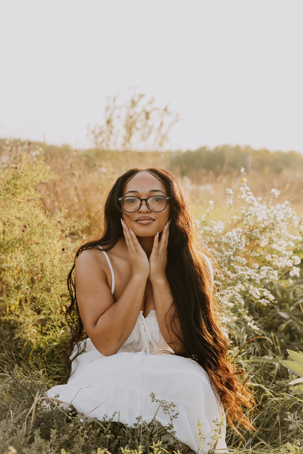 Woman with long dark hair and glasses in white dress sitting among wildflowers in a sunlit field.