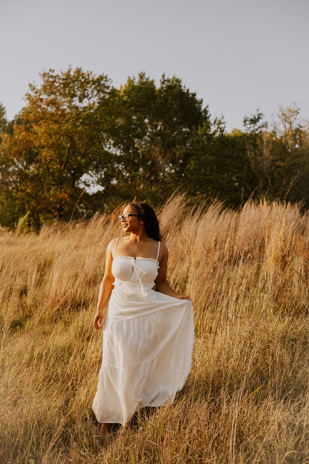 Woman in white sundress walking through golden field with trees in background during golden hour.