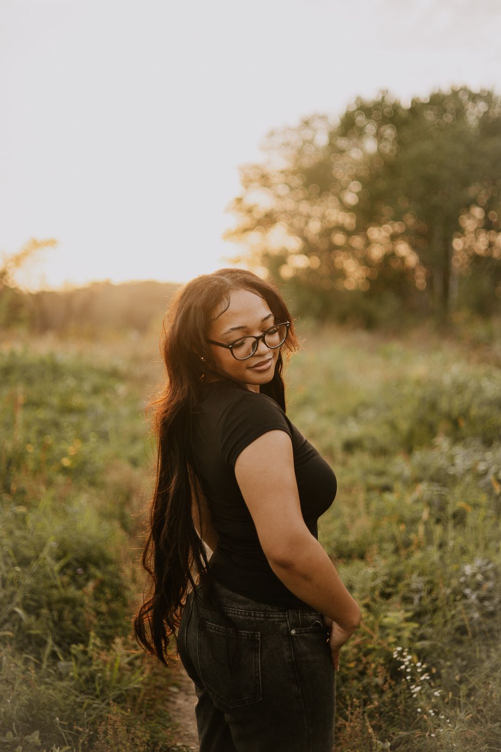 Young woman with long dark hair and glasses smiling over shoulder in golden hour field.