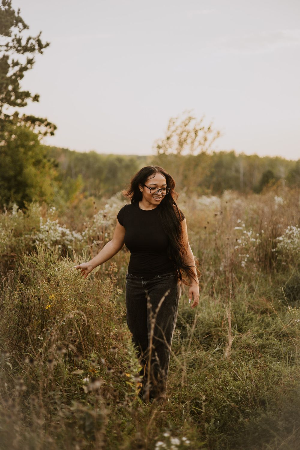 Woman in black top and jeans walking through a wildflower meadow at golden hour, smiling softly.