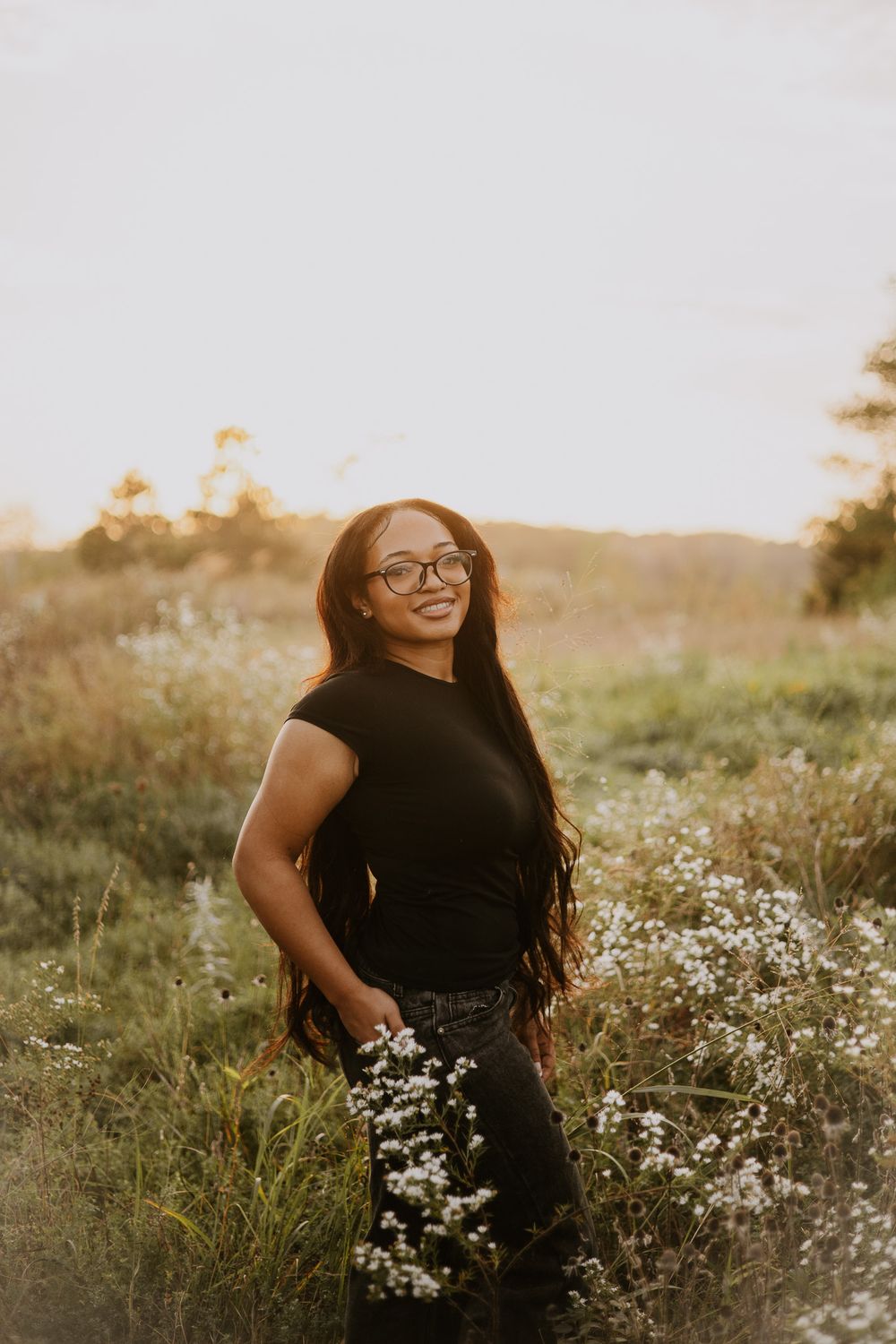 Young woman with long hair and glasses smiles in a sunlit field holding white wildflowers at golden hour.