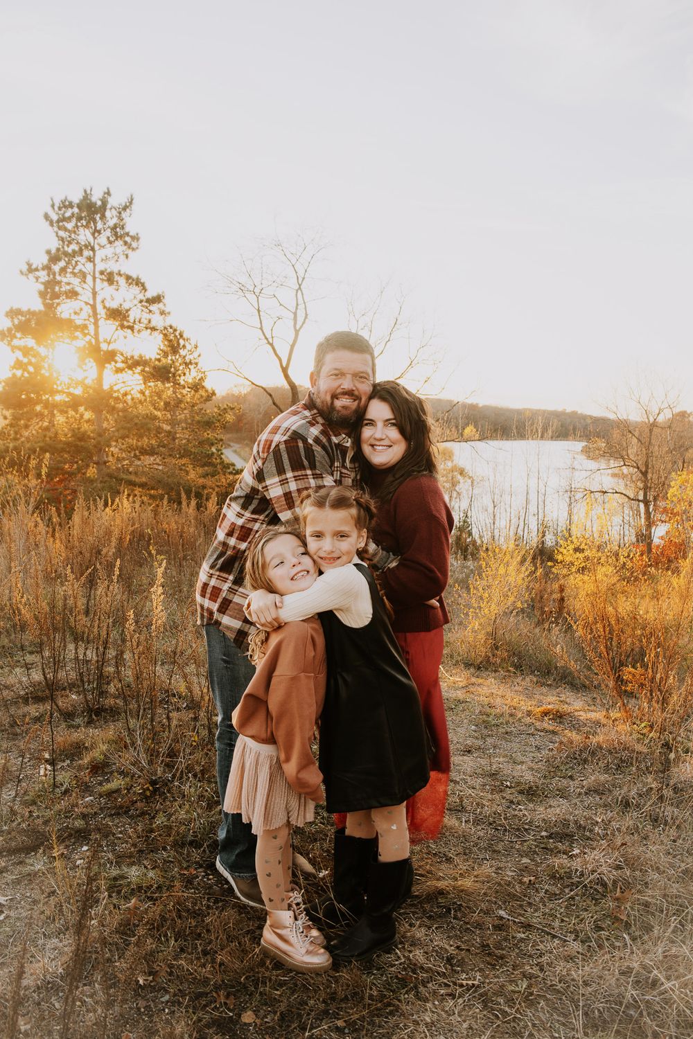 Family of four hugging outdoors at golden hour near a lake, surrounded by autumn foliage and tall grasses.