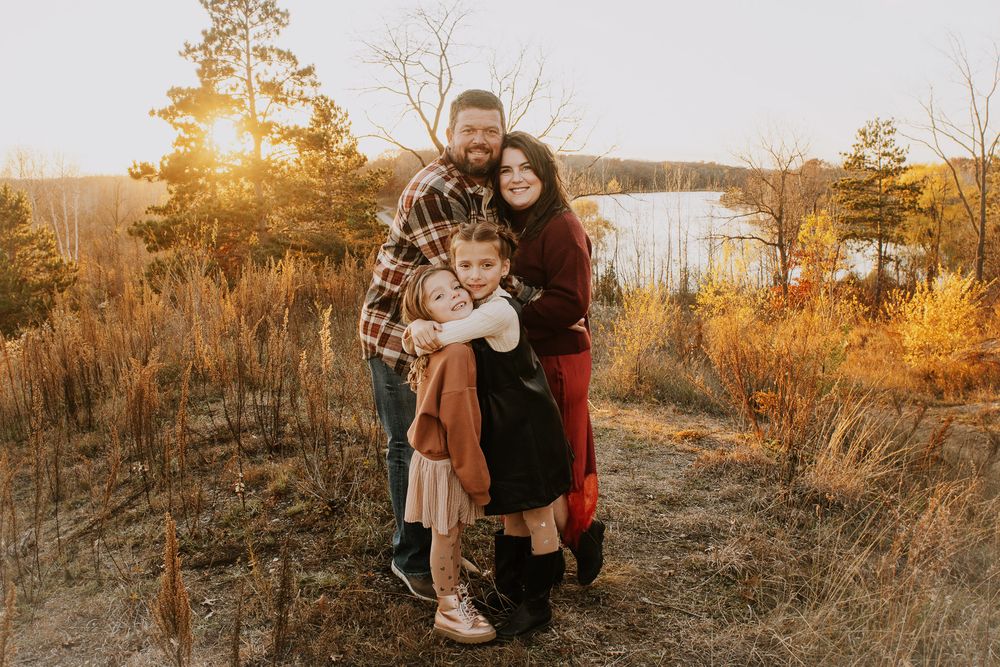Family of four posing outdoors at golden hour near a lake surrounded by autumn foliage.