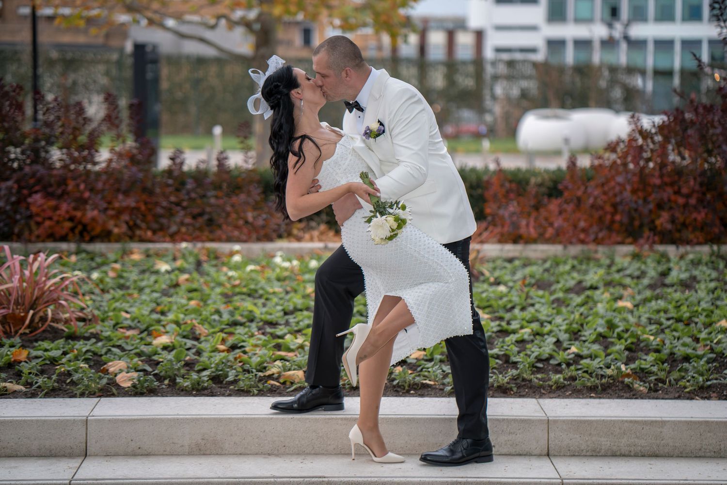 Bride and groom share a romantic dip kiss outdoors in wedding attire, white dress and tuxedo, holding bouquet.