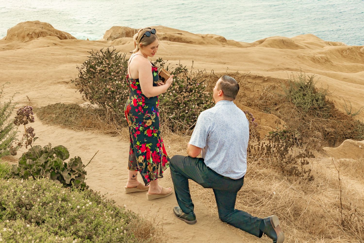 Man proposing on one knee to woman in floral dress on scenic coastal cliffside path overlooking the ocean.