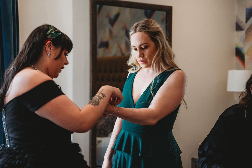 Bride in black dress helping bridesmaid in green gown with outfit adjustment.