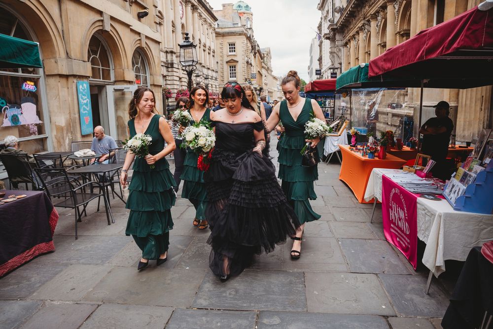 Bride in black tiered gown walks with bridesmaids in emerald green dresses through city street.