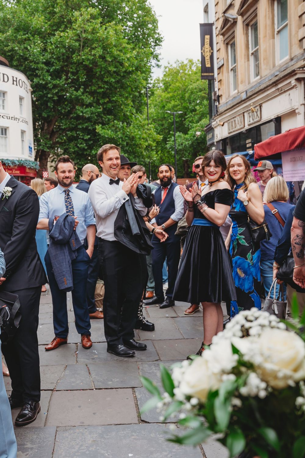 Wedding guests clap and celebrate outside a historic building on a sunny day.