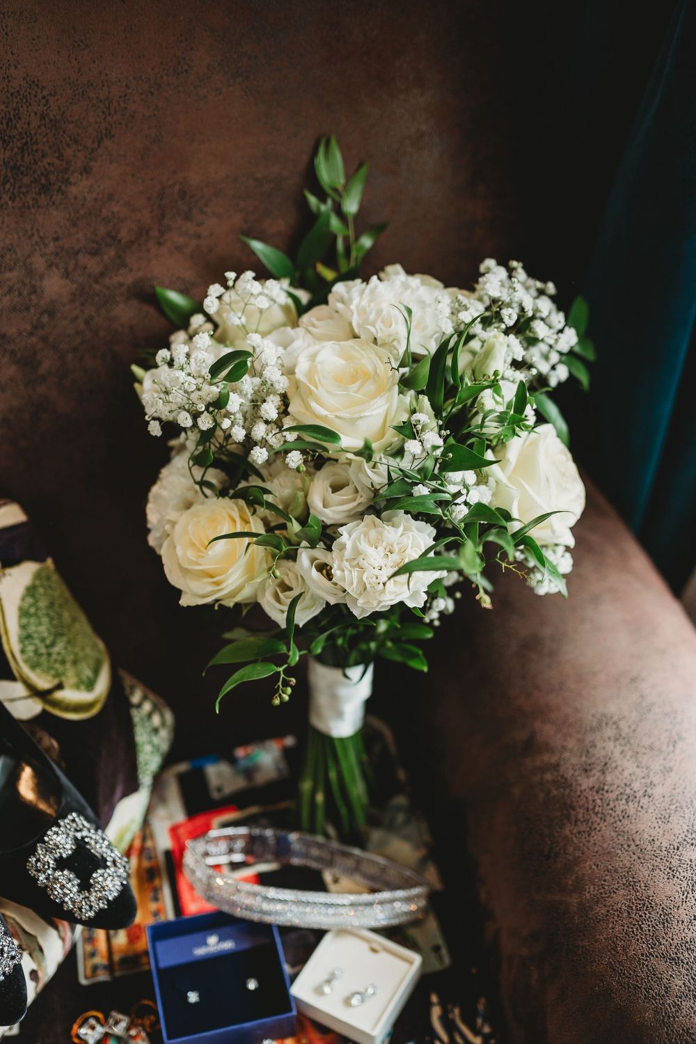 Bridal bouquet of white roses and baby's breath rests beside jewelry and wedding accessories.