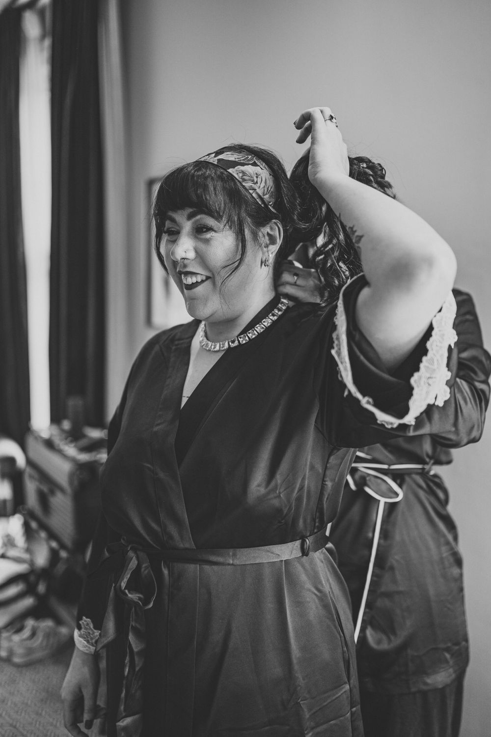 Bride laughing joyfully in black robe while getting hair styled during wedding preparations.