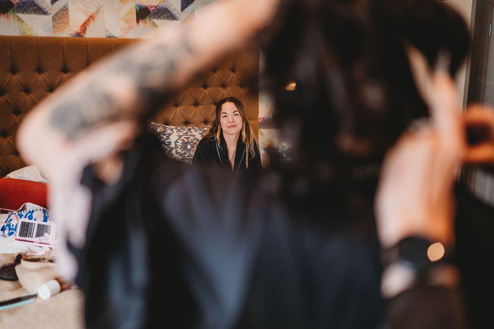 Bride with tattoos getting hair done, reflected in mirror during wedding day preparations.