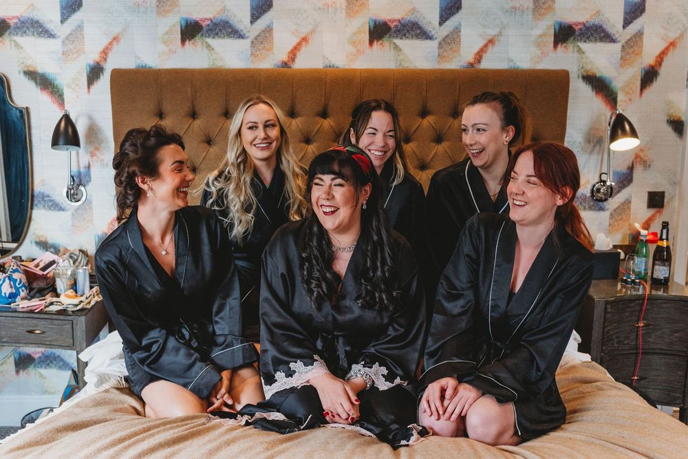 Bridesmaids in black satin robes laughing together on a bed during wedding morning preparations.