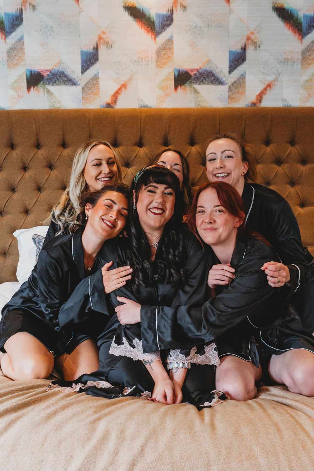 Six bridesmaids in black satin robes laugh and cuddle together on a hotel bed before the wedding.