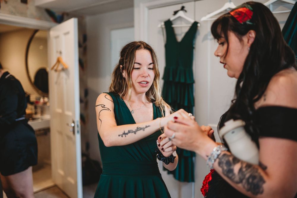 Bridesmaid in emerald green dress helps another with jewelry during wedding preparations in bright dressing room.